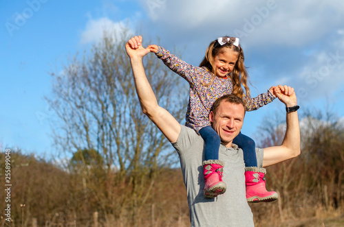 Young father carrying his adorable daughter on his shoulders on a sunny day in the nature. Family bonding and happiness.