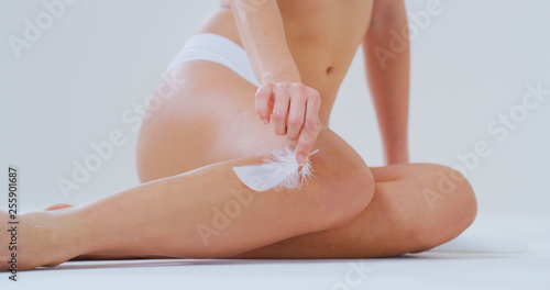 Photos Close up of young woman with perfect body touching gently her hairless soft and silky legs with white feather after depilation isolated on a white background
