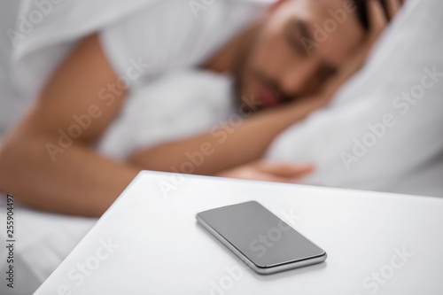 technology and people concept - close up of smartphone on bedside table near young man sleeping in bed at home in morning