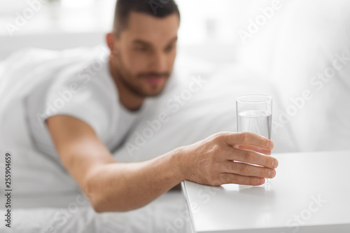 morning and hydration concept - close up of thirsty man reaching to glass of water on bedside table from bed at home