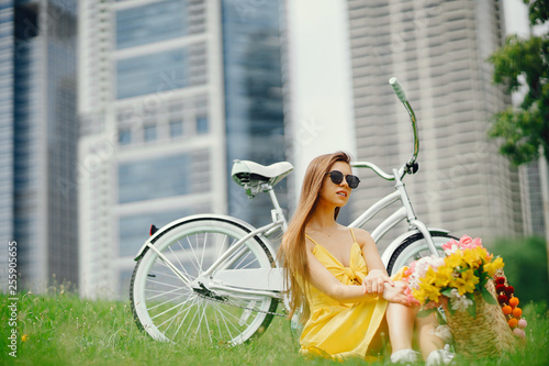 a cute and stylish girl in a yellow dress and long hair sitting on agrass near bicycle with a basket of flowers in the sunny summer city photo