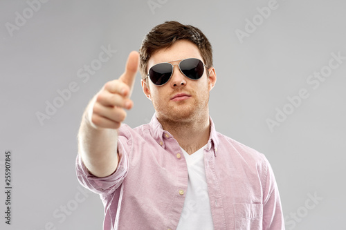 summer accessories, style and people concept - young man in shirt and black aviator sunglasses making hand gun gesture over grey background