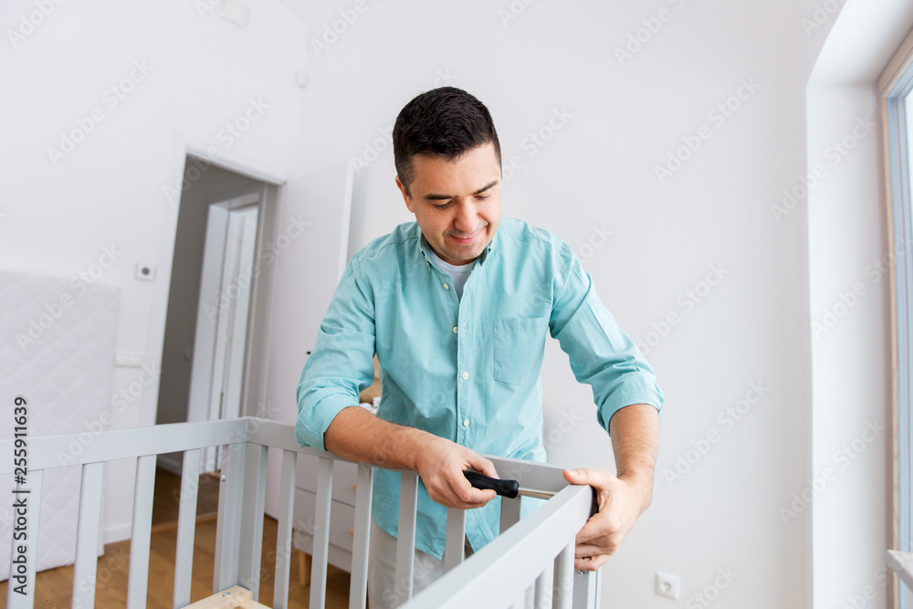 parenthood, fatherhood and nursery concept - happy middle-aged father with screwdriver assembling baby bed at home
