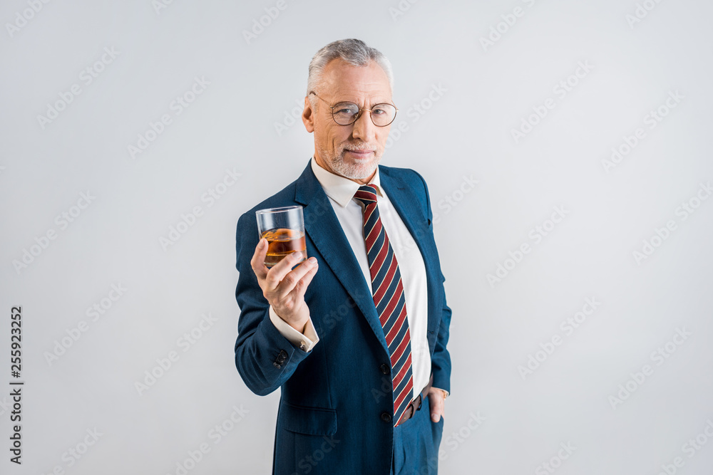 cheerful mature businessman holding glass of whiskey while standing with hand in pocket isolated on grey cheerful mature businessman holding glass of whiskey while standing with hand in pocket isolated on grey