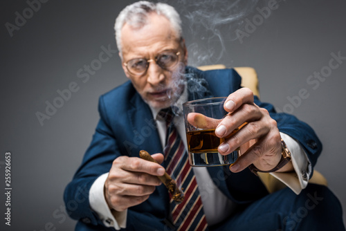 selective focus of mature businessman in suit holding glass of whiskey and cigar on grey