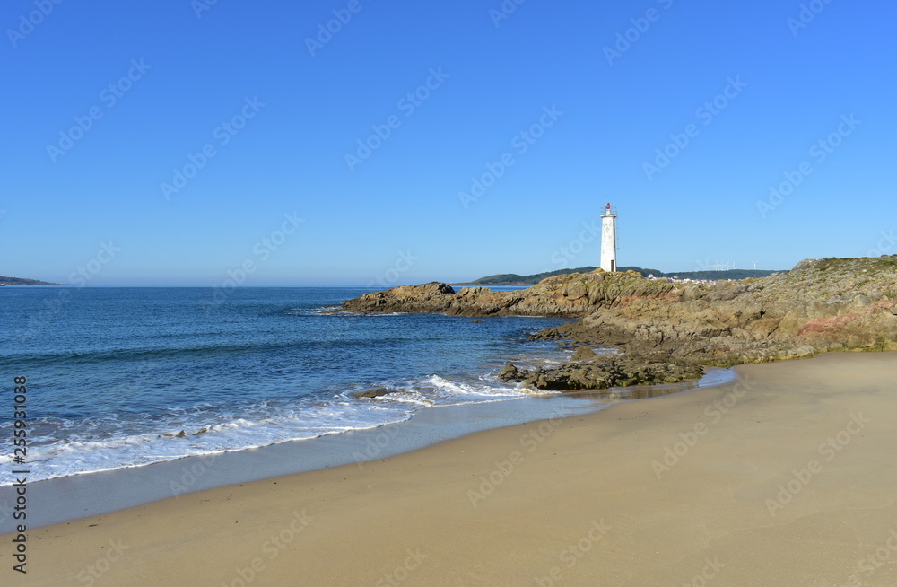 Beach with white lighthouse on the rocks and blue sea with foam. Sunny day, Galicia, Spain.