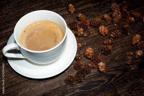 white cup of coffee on wood table and some pine cones on the background.