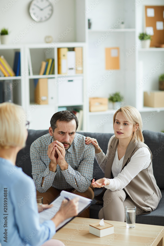 Blonde young woman explaining something to counselor while sitting on couch next to her husband Blonde young woman explaining something to counselor while sitting on couch next to her husband