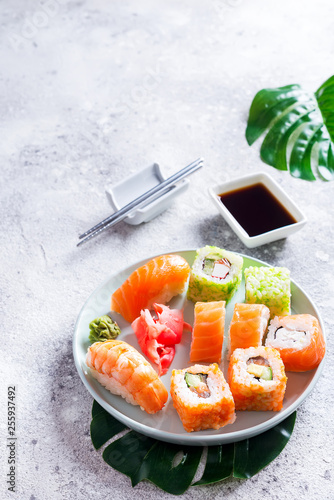 Various sushi on ceramic plate with metal Korean sticks on light stone background with green leaves, flat lay