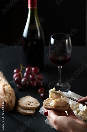 glass of red wine with a bottle, cluster of grapes, women hand placing brie cheese to rustick piece of bread  with antique knife. photo