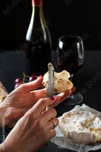 glass of red wine with a bottle, cluster of grapes, women hand placing brie cheese to rustick piece of bread  with antique knife. photo