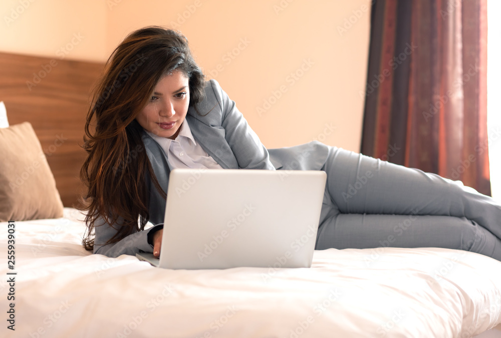 Young businesswoman working from hotel room on business trip, woman lying on the bed and using laptop. Young businesswoman working from hotel room on business trip, woman lying on the bed and using laptop.