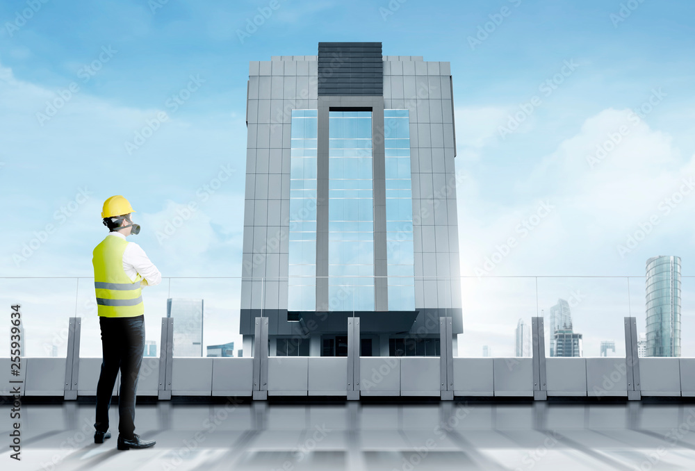 Rear view of asian worker man in safety vest, gloves, yellow helmet and protective mask standing on modern terrace and looking at skyscrapers