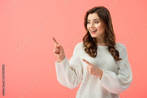 smiling and beautiful woman in white sweater pointing with fingers and looking away isolated on pink
