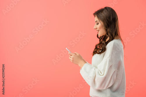 smiling and attractive woman in white sweater holding smartphone isolated on pink