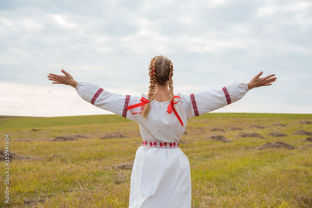 A girl in a Slavic dress with a red ribbon in her hair is standing in a field with her arms open to the sky A girl in a Slavic dress with a red ribbon in her hair is standing in a field with her arms open to the sky