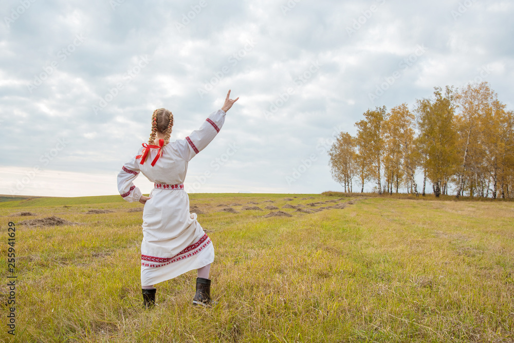 A girl in a Slavic dress with a red ribbon in her hair is standing in a field with her arms open to the sky A girl in a Slavic dress with a red ribbon in her hair is standing in a field with her arms open to the sky
