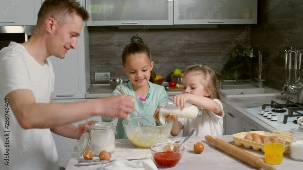 Two adorable primary school girls and their father making dough in kitchen together, one of girls using whisk and one pouring milk into bowl, father talking to them Two adorable primary school girls and their father making dough in kitchen together, one of girls using whisk and one pouring milk into bowl, father talking to them