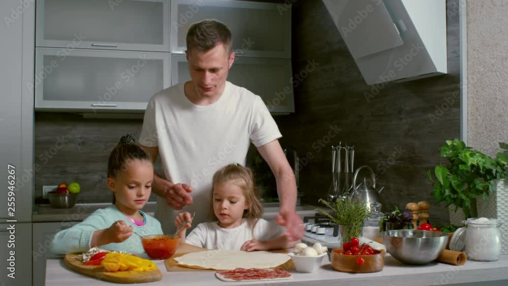 Young father and two daughters making pizza in kitchen together, man teaching girls to spread tomato sauce on dough Young father and two daughters making pizza in kitchen together, man teaching girls to spread tomato sauce on dough