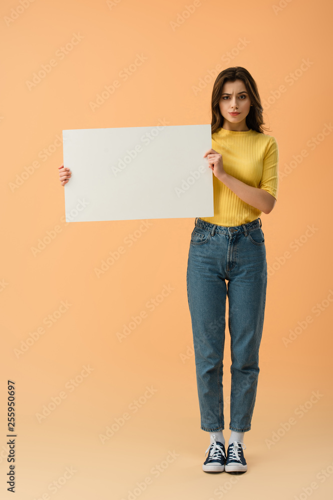 Pensive brunette girl in jeans and jumper holding blank placard on orange background Pensive brunette girl in jeans and jumper holding blank placard on orange background