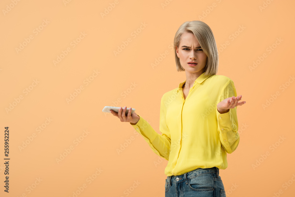 Dissatisfied blonde woman in yellow shirt holding smartphone isolated on orange Dissatisfied blonde woman in yellow shirt holding smartphone isolated on orange