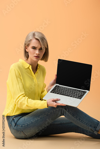 Upset blonde woman in yellow shirt showing laptop with blank screen on orange background