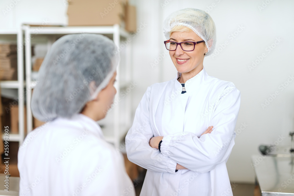 Blonde Caucasian employee in sterile uniform talking to supervisor about quality of products and holding arms crossed. Food factory interior.
