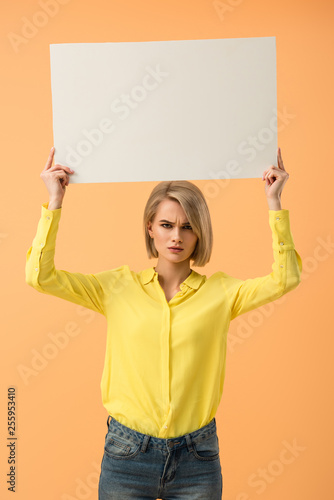Dissatisfied blonde girl in yellow shirt holding blank placard isolated on orange