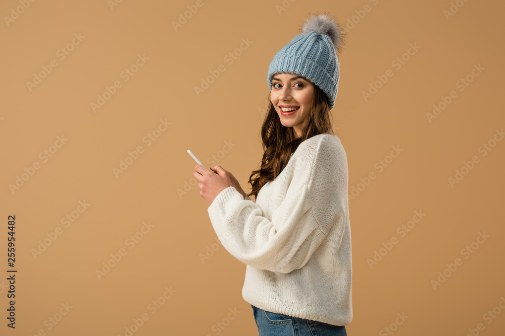 Excited curly woman in knitted hat holding smartphone isolated on beige