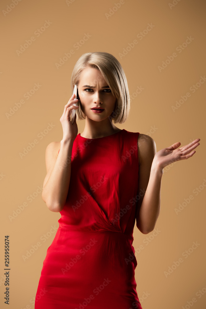 Upset blonde girl in red dress talking on smartphone isolated on beige Upset blonde girl in red dress talking on smartphone isolated on beige