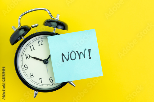 Sticky post with handwriting the word Now stick on alarm clock on solid yellow background with copy space using as stop procrastination, self discipline or motivation to complete task
