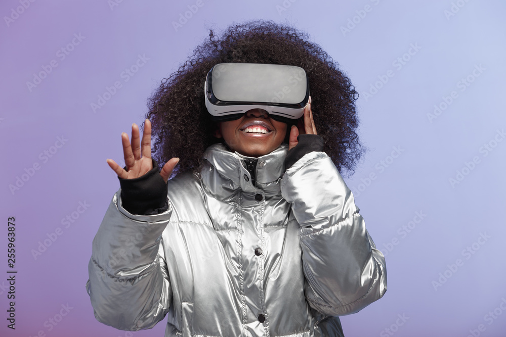 Modern curly brown-haired girl dressed in a silver-colored jacket uses the virtual reality glasses poses in the studio on neon background Modern curly brown-haired girl dressed in a silver-colored jacket uses the virtual reality glasses poses in the studio on neon background