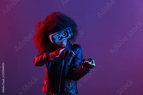 Curly dark haired girl dressed in black leather jacket and gloves uses the virtual reality glasses on her head in the dark studio with neon light