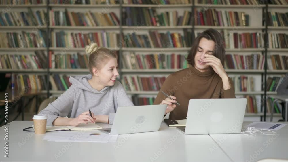 Pan shot of two young students male with long hair dressed in brown sweatshirt and female in grey hoodie talking in the library. Two students with laptops sitting side by side working on project Pan shot of two young students male with long hair dressed in brown sweatshirt and female in grey hoodie talking in the library. Two students with laptops sitting side by side working on project