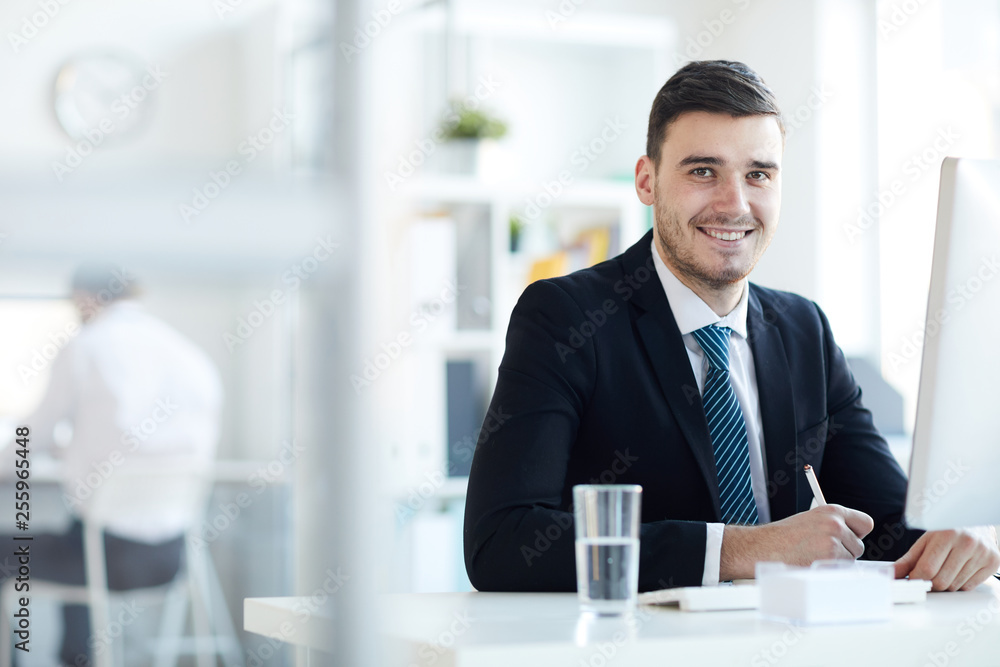Young successful businessman in formalwear sitting by desk in front of computer monitor and organizing work Young successful businessman in formalwear sitting by desk in front of computer monitor and organizing work
