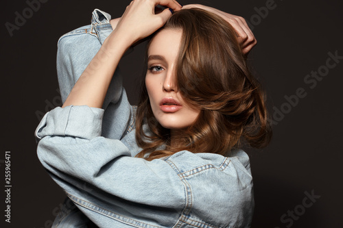 Beautiful brunette girl with long flowing hair dressed in jeans jacket poses holding her hands on her head on the dark background in the studio