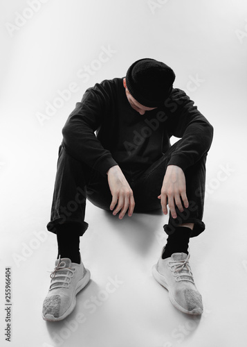 Guy dressed in black jeans, sweatshirt, hat and gray sneakers is sitting on the floor in the studio on the white background