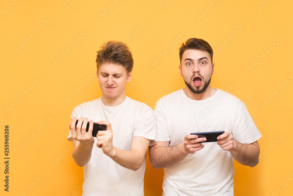 Emotional gamers play mobile games on a smartphone, wear white t-shirts, one looks at the camera, the other in the smartphone screen, they are isolated on a yellow background. Emotional gamers play mobile games on a smartphone, wear white t-shirts, one looks at the camera, the other in the smartphone screen, they are isolated on a yellow background.