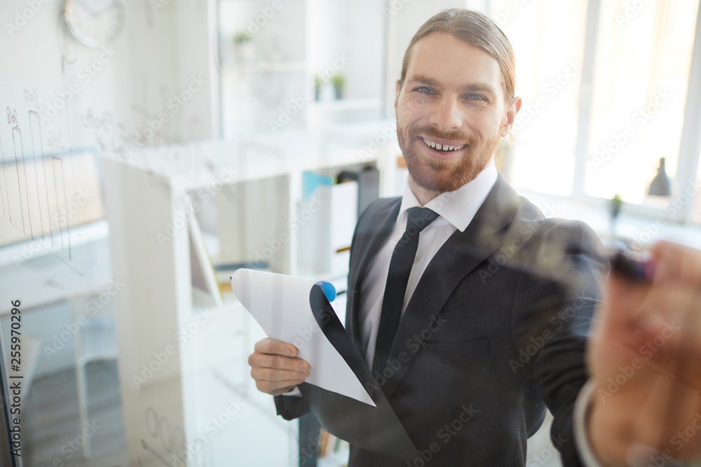 Happy young smiling analyst in suit standing by board while working with financial data or making presentation