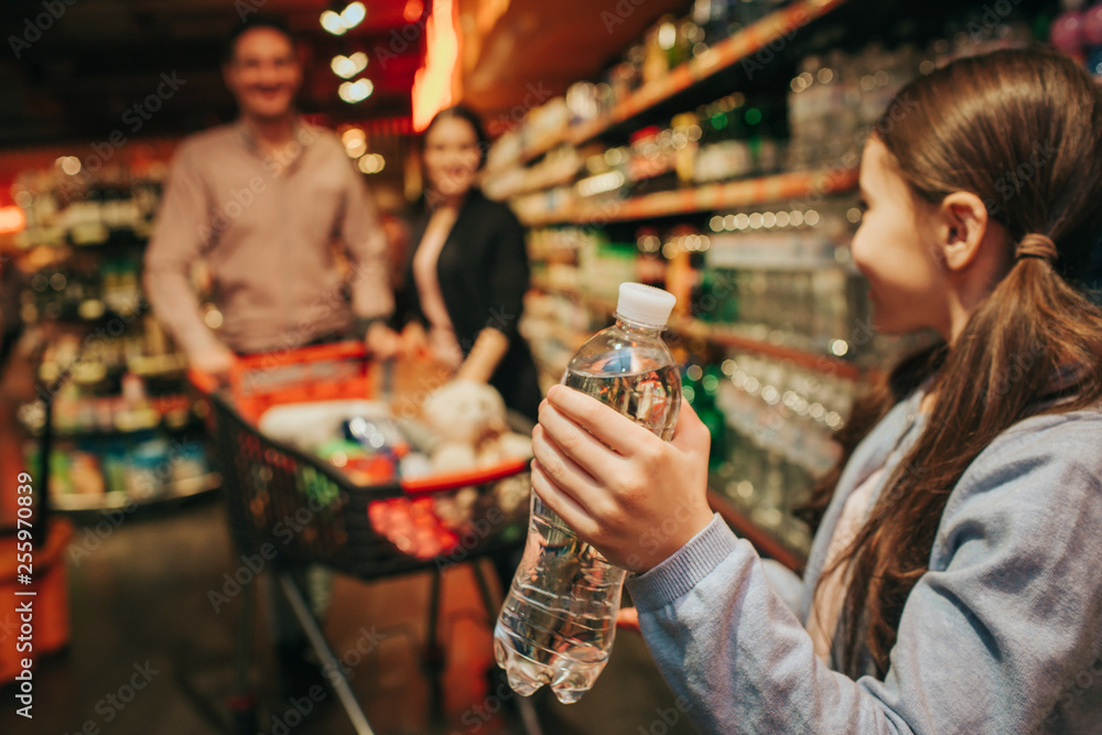 Young parents and daughter in grocery store. Little girl hold water bottle in hands and look at parents. They stand behind and carry grocery trolley.