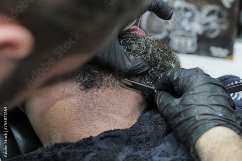 Barber shaves the beard of an elderly man with gray hair sharp razor