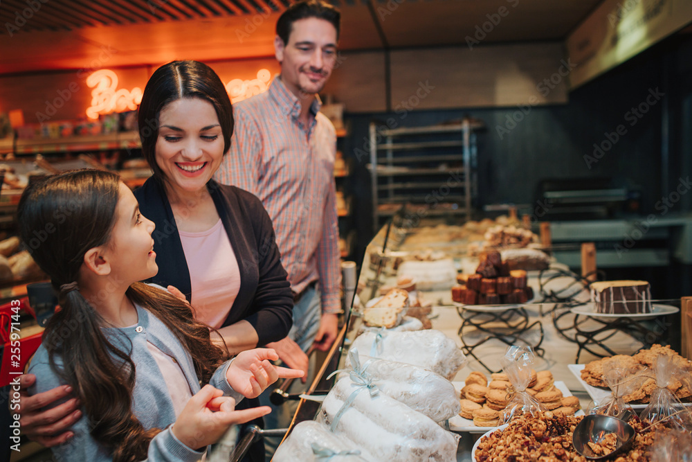 Young parents and daughter in grocery store. Girl look at mother with happiness. Woman smile. Man stand behind and look at them.