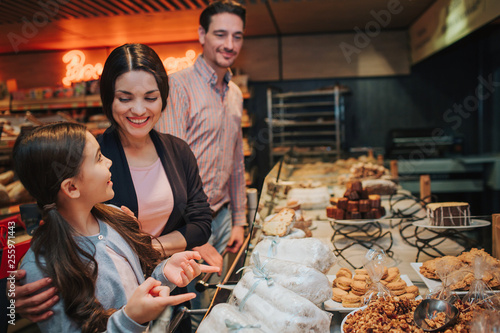 Young parents and daughter in grocery store. Girl look at mother with happiness. Woman smile. Man stand behind and look at them.