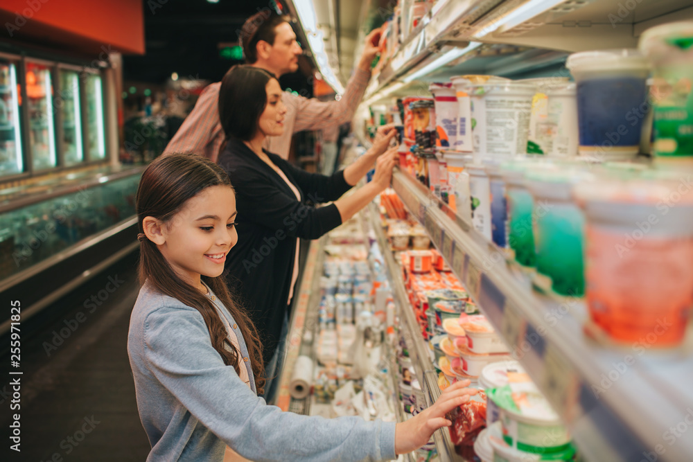 Young parents and daughter in grocery store. They stand at dairy shelf and choosing. Girl touch jars and look down. Parents stand behind.