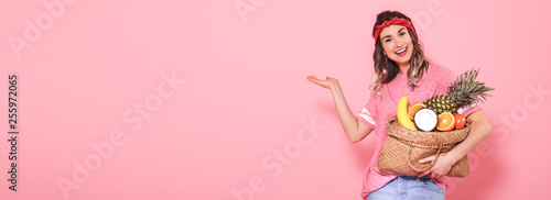 Beautiful young girl in pink t-shirt, holds a full straw bag of fruit on pink background