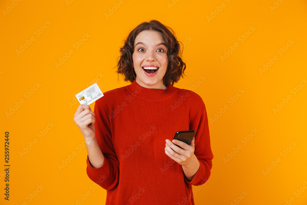 Portrait of feminine woman using cell phone and credit card while standing isolated over yellow background Portrait of feminine woman using cell phone and credit card while standing isolated over yellow background