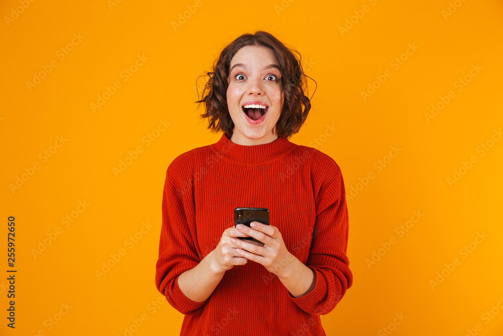Fototapeta premium Portrait of caucasian woman holding and using cell phone while standing isolated over yellow background