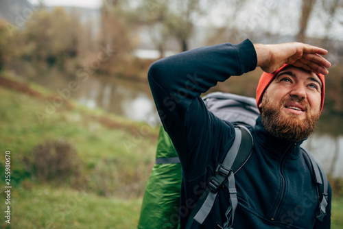 Explorer young man hiking in mountains with travel backpack looking to a beautiful view with hand on the forehead. Traveler bearded man relaxing after trekking. Travel, people and lifestyle concept