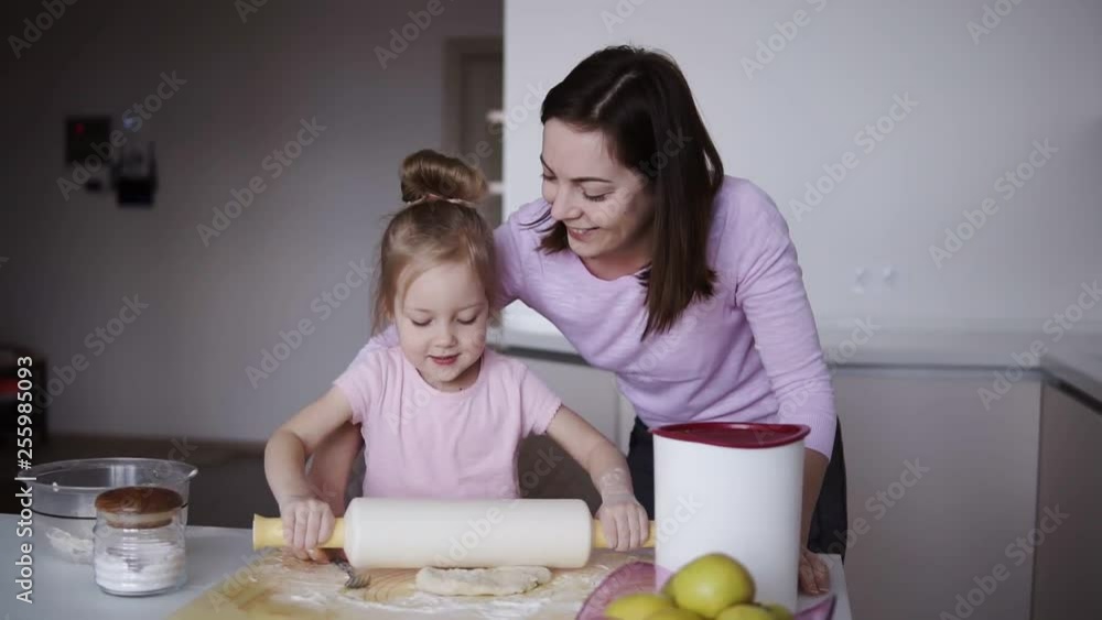 Beautiful young loving mother teaching her cute little daughter rolling out dough with a rolling pin while cooking together at the kitchen. Cooking and having fun Beautiful young loving mother teaching her cute little daughter rolling out dough with a rolling pin while cooking together at the kitchen. Cooking and having fun