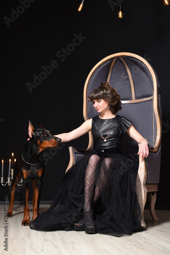 A girl in a black dress is sitting in a big gray chair and next to her is a Doberman dog, she touches him © pridannikov
